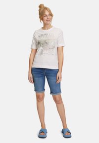 T-shirt bianca in cotone con stampa grafica, maniche corte, abbinata a pantaloni corti di denim arrotolati e sandali blu a scivolo con superficie texturizzata.