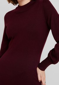 Burgundin värinen neulemekko, jossa on korkea kaulus, pitkät hihat ja ribatettua pintaa. Slim fit -muotoilu, jossa on kerättyjä yksityiskohtia hihansuissa.