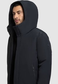 Černá péřová bunda s vysokým límcem a velkou kapucí, se zipovou kapsou na boku a minimalistickými designovými detaily.