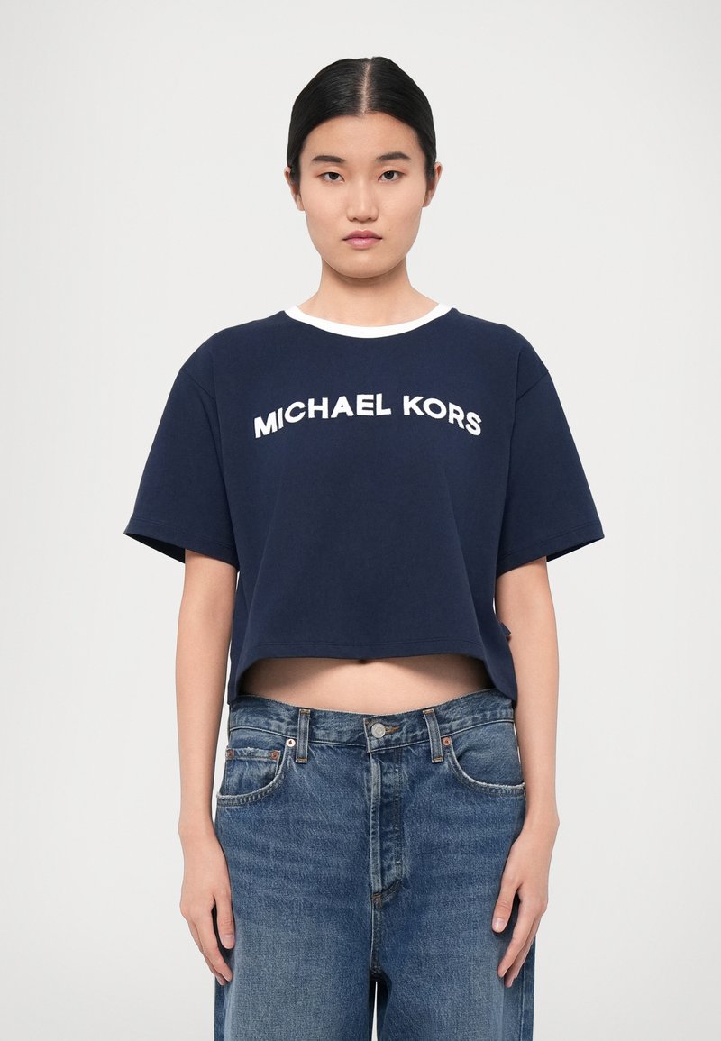 Navy sininen lyhyt t-paita, jossa on valkoinen "MICHAEL KORS" logo, lyhyet hihat ja pyöreä pääntie. Yhdistetty vaaleansiniset farkut.