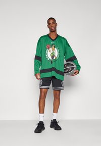 Zöld, hosszú ujjú póló fekete részletekkel, Boston Celtics logóval és grafikával. Fekete sorttal és kosárlabdával párosítva.