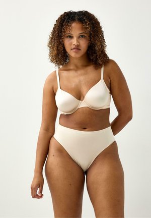 Kvinde med krøllet hår iført beige bh og høj talje trusser, står foran en ensfarvet hvid baggrund.