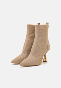 Botas hasta la rodilla con punta afilada en tejido dorado texturizado y tacón en forma de cono metálico. Presentan un ajuste ceñido y una lengüeta en la parte posterior.