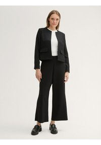 Chaqueta negra texturizada corta con dos bolsillos frontales, llevada sobre una blusa blanca de manga larga y combinada con pantalones negros de pierna ancha y mocasines negros.