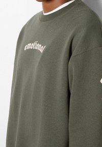 Olivgrüner Sweatshirt aus weichem Material, mit dem Wort "emotional" in einer hellroten, geschwungenen Schriftgröße über der Brust.