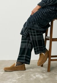 Brune suède slippers met bontvoering, grijze geribde badjas en zwart geruite broek, zittend op een houten stoel tegen een effen achtergrond.