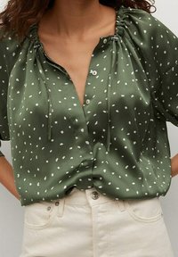 Blusa verde con pois bianchi, realizzata in un materiale setoso. Presenta un scollo a V, dettagli con cordoncino in corrispondenza del collo e una vestibilità morbida.