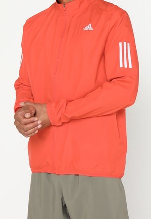 Homme portant une veste de sport Adidas orange vif avec des bandes blanches sur les manches et un logo sur la poitrine, associée à un pantalon gris clair.