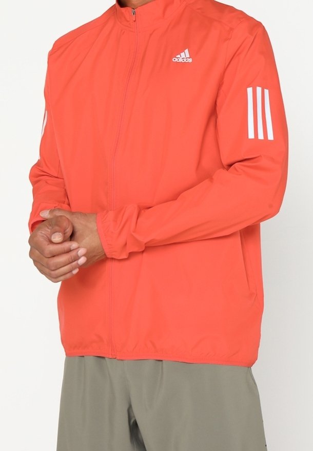 Homme portant une veste de sport Adidas orange vif avec des bandes blanches sur les manches et un logo sur la poitrine, associée à un pantalon gris clair.