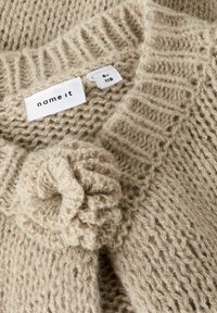 Gilet en tricot beige avec un motif texturé, doté d'une fermeture décorative à pompon. Les étiquettes indiquent la taille 6 ans et la marque "name it".
