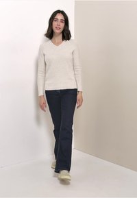Hellbeige V-Ausschnitt-Pullover mit strukturierter Strickoberfläche, kombiniert mit dunkelblauen Schlagjeans. Das Model trägt helle Sneaker.