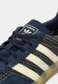 Σκούρο μπλε αθλητικό παπούτσι με σουέντ υλικό και διακριτικά από δίχτυ, με τρεις λευκές ρίγες, δετά κορδόνια και λογότυπο στη γλώσσα.