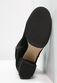 Bottine noire avec une semelle en caoutchouc texturée et un talon bloc, vue par le dessous avec un détail de fermeture éclair latérale visible.