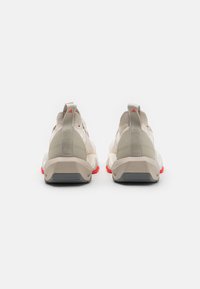 Beige athletic sneakers with a textured upper, red and gray accents, and a cushioned sole. Features a pull tab and branding on the heel.