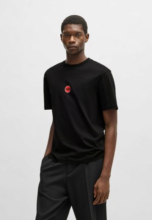 Black crew-neck t-shirt made of cotton, featuring a red circular logo on the chest. Short sleeves and relaxed fit. Worn with dark trousers.