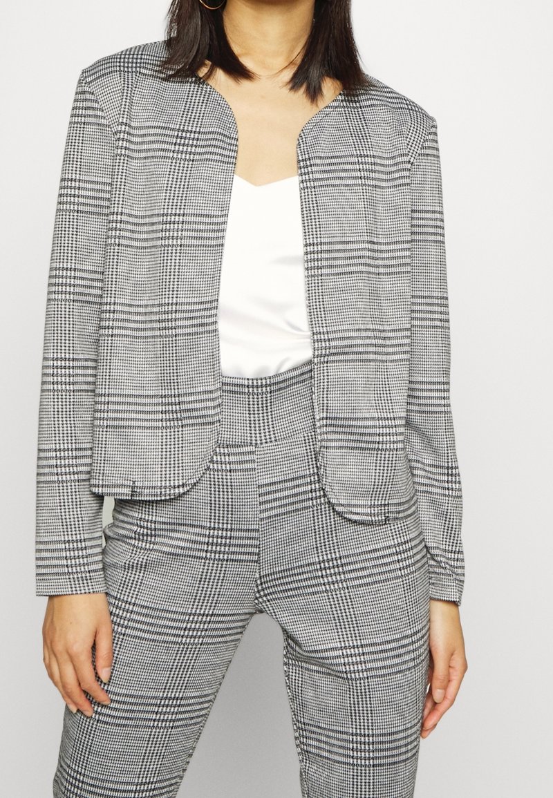 Blazer à carreaux noir et blanc avec un devant court et ouvert, un col arrondi et des manches longues, associé à un pantalon assorti à carreaux.