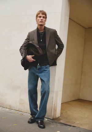 Młody mężczyzna stoi przy beżowej ścianie, ubrany w kratkę marynarkę, czarną koszulę, luźne niebieskie dżinsy, czarne buty, trzyma ciemnobrązową torbę.