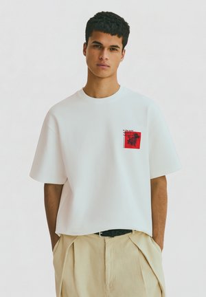 Ung mand iført hvid oversized t-shirt med lille rød grafik og tekst på brystet, beige bukser, står med hænderne i lommerne.