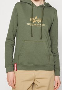 Felpa verde realizzata in tessuto morbido, con una stampa del logo dorato, coulisse regolabile, tasca frontale e un'etichetta rossa con logo sul lato.