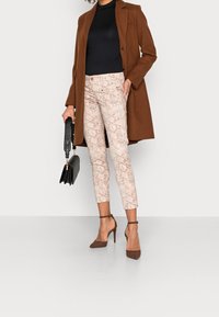 Hnědý vlněný kabát přes černý rolák, doplněný růžovými kalhotami se zebrovaným potiskem a hnědými vysokými podpatky. Drží černou kabelku.