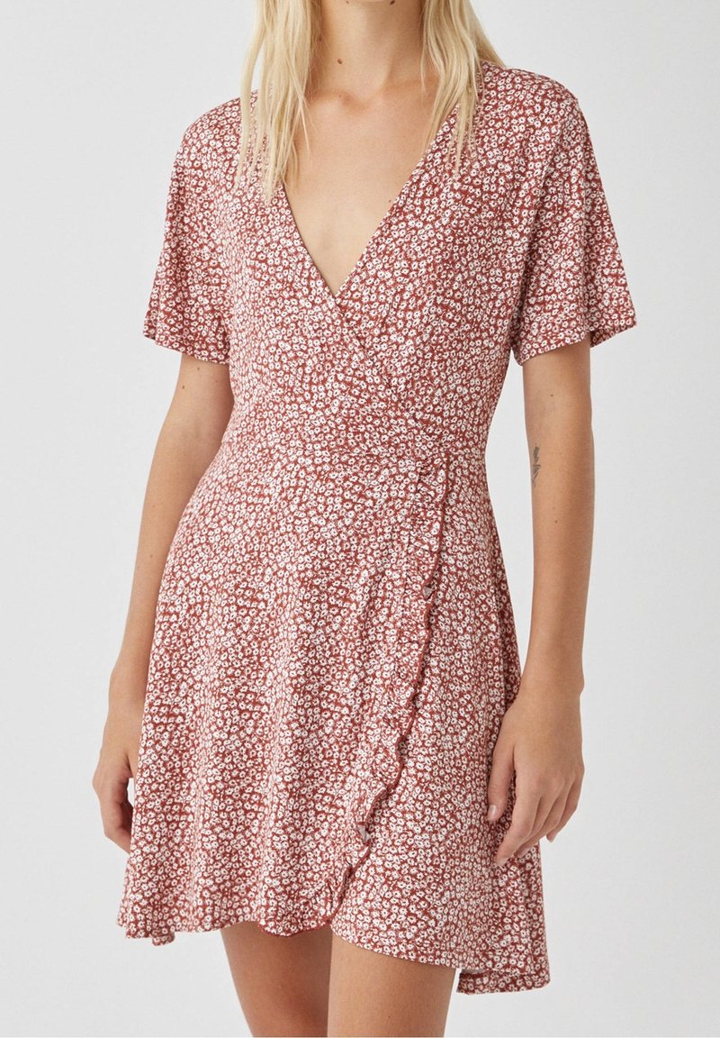 Femme portant une robe portefeuille rouge à manches courtes avec un petit imprimé floral blanc et un ourlet à volants sur un fond uni.