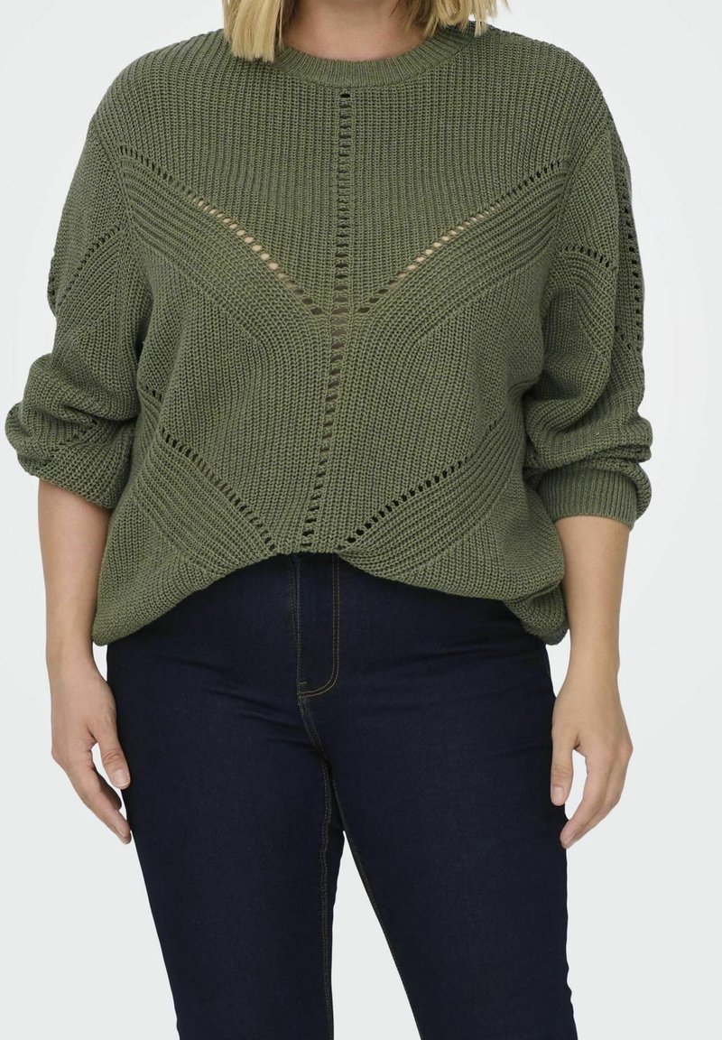 Personne portant un pull en maille vert olive avec des motifs géométriques en dentelle et un jean bleu foncé, mains détendues le long du corps.