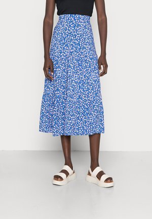 Person iført en blå blomster midi nederdel kombineret med en sort top og hvide plateau sandaler står på et lyst gulv.