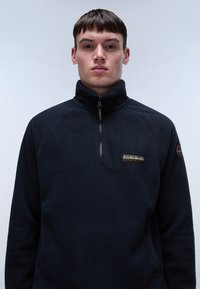 Pullover in pile nero con colletto alto, zip e toppa con logo sul petto. Presenta una texture morbida e maniche raglan.