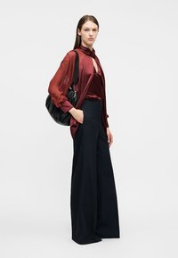 Jauna sieviete valkā vīnogu krāsas satīna blūzi, caurspīdīgu vīnogu krāsas virsblūzi, plašas melnas bikses un nēsā melnu plecu somu.