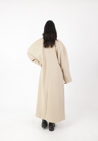 Cappotto lungo beige con spalle drop, maniche ampie e silhouette dritta. Tessuto liscio con una cucitura centrale che scorre lungo la schiena.