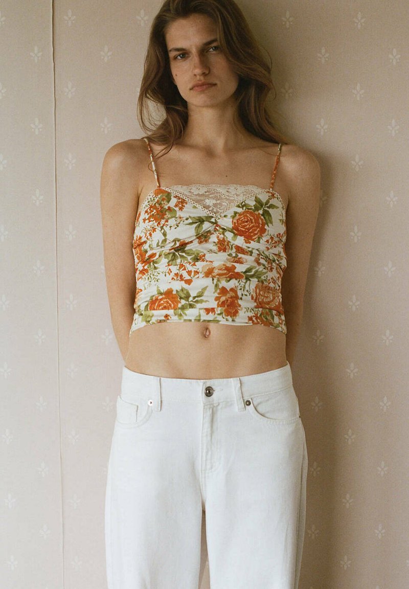 Jeune femme debout contre un mur beige à motifs, portant un jean blanc taille haute et un caraco court fleuri avec bordure en dentelle.