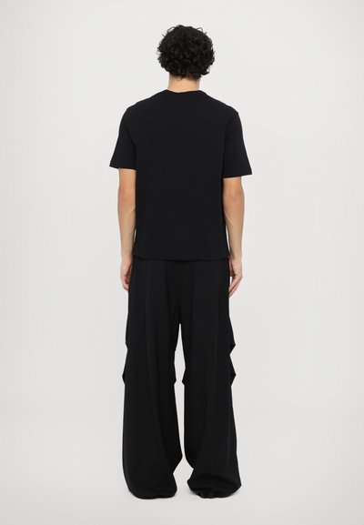 Czarny t-shirt z krótkim rękawem noszony z szerokimi czarnymi spodniami, które mają warstwowe boki, prezentując luźny krój i miękką teksturę materiału.