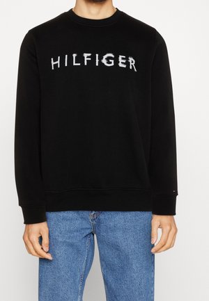 Mężczyzna ubrany w czarną bluzę z długim rękawem z napisem "HILFIGER" na piersi, w niebieskich dżinsach, stojący na białym tle.