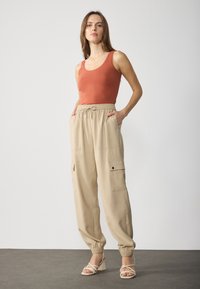 Beige cargo pants with elastic waistband and side pockets, paired with a rust-colored tank top, set against a plain backdrop.