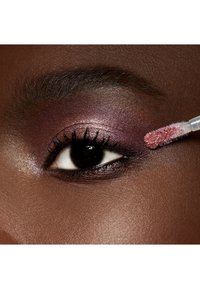 Un fard à paupières liquide dans des teintes de violet et de rose est appliqué sur la paupière, mettant en valeur l'œil avec des paillettes et un eyeliner bien défini.