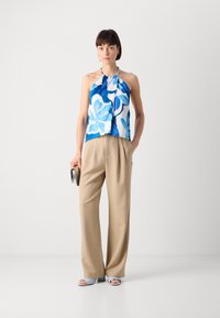 Haut à bretelles à motifs floraux bleu et blanc, associé à un pantalon taille haute beige. Les accessoires incluent une pochette argentée et des sandales bleu clair.
