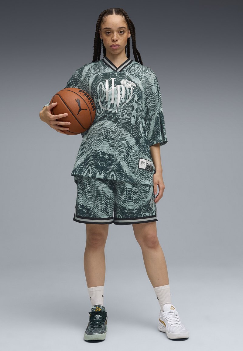 Green patterned oversized jersey with snake design, paired with matching shorts. Player holds a brown basketball, wearing black and white sneakers.