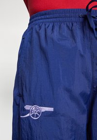 Lehké šortky námořnicky modré barvy s texturovaným povrchem, s vyšívaným logem děla světle fialové barvy na levé noze a elastickým pasem.