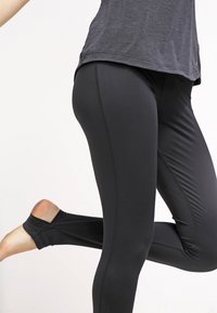 Černé legíny vyrobené z elastického materiálu, s přiléhavým střihem a bezešvým šitím, s detailem výřezu na kotníku.