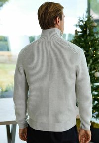 Pull gris clair à côtes avec un col haut, des manches longues et un motif de tricot texturé, présentant une taille et des poignets ajustés.