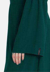 Îmbrăcăminte verde, cu mâneci lungi și manșete în formă de clopot. Textile moi cu un luciu subtil. Etichetă neagră cu logo alb brodat pe manșetă.