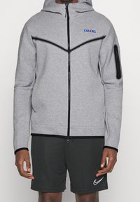 Sudadera gris con cremallera, capucha, acentos en negro y un logo en azul. Textura suave, diseño ajustado y bolsillo lateral con cremallera. Combinada con pantalones cortos oscuros.