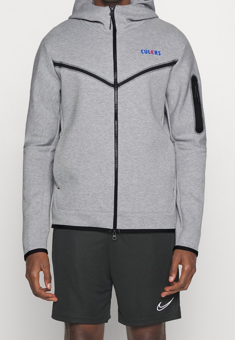 Sudadera gris con cremallera, capucha, acentos en negro y un logo en azul. Textura suave, diseño ajustado y bolsillo lateral con cremallera. Combinada con pantalones cortos oscuros.