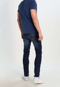 Dunkelblaue Skinny Jeans mit einer verblassten Textur, kombiniert mit einem navy-blauen T-Shirt mit kurzen Ärmeln und weißen Sneakers, von hinten betrachtet.