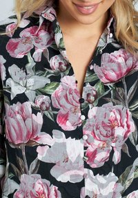 La camicia a maniche corte presenta un fondo nero, grandi fiori rosa e bianchi, con una consistenza morbida e piccoli bottoni bianchi lungo la chiusura.