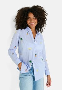 Camisa a rayas azul con diseños bordados, que incluyen piñas, palmeras, arcoíris y motivos de sol. Manga larga y cierre de botones en el frente.