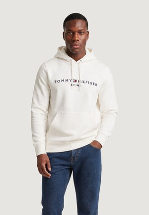 Mand iført en hvid Tommy Hilfiger hættetrøje med logo og blå jeans, stående med den ene hånd i lommen mod en ensfarvet baggrund.