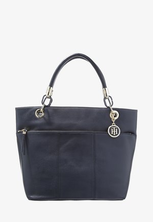 Czarna skórzana torba typu tote ze złotymi elementami, zewnętrzną kieszenią na zamek, podwójnymi uchwytami oraz okrągłą złotą zawieszką z inicjałami wiszącą po prawej stronie.