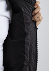 Black jacket interior featuring smooth, matte fabric texture with a zip closure and a side pocket, held open with a hand.