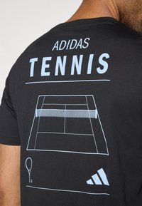 Melna kokvilnas T-krekls ar gaiši ziliem grafiskajiem attēliem, kuros attēlots tenisa laukums, vārds "ADIDAS" un tenisa rakete logo tuvu apakšai.